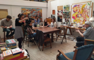 Joyce Tsai's class visits Mel Andringa in Cedar Rapids. He is seated in an art studio with some colorful paintings. Students sit around a table while Andringa talks about Intermedia.